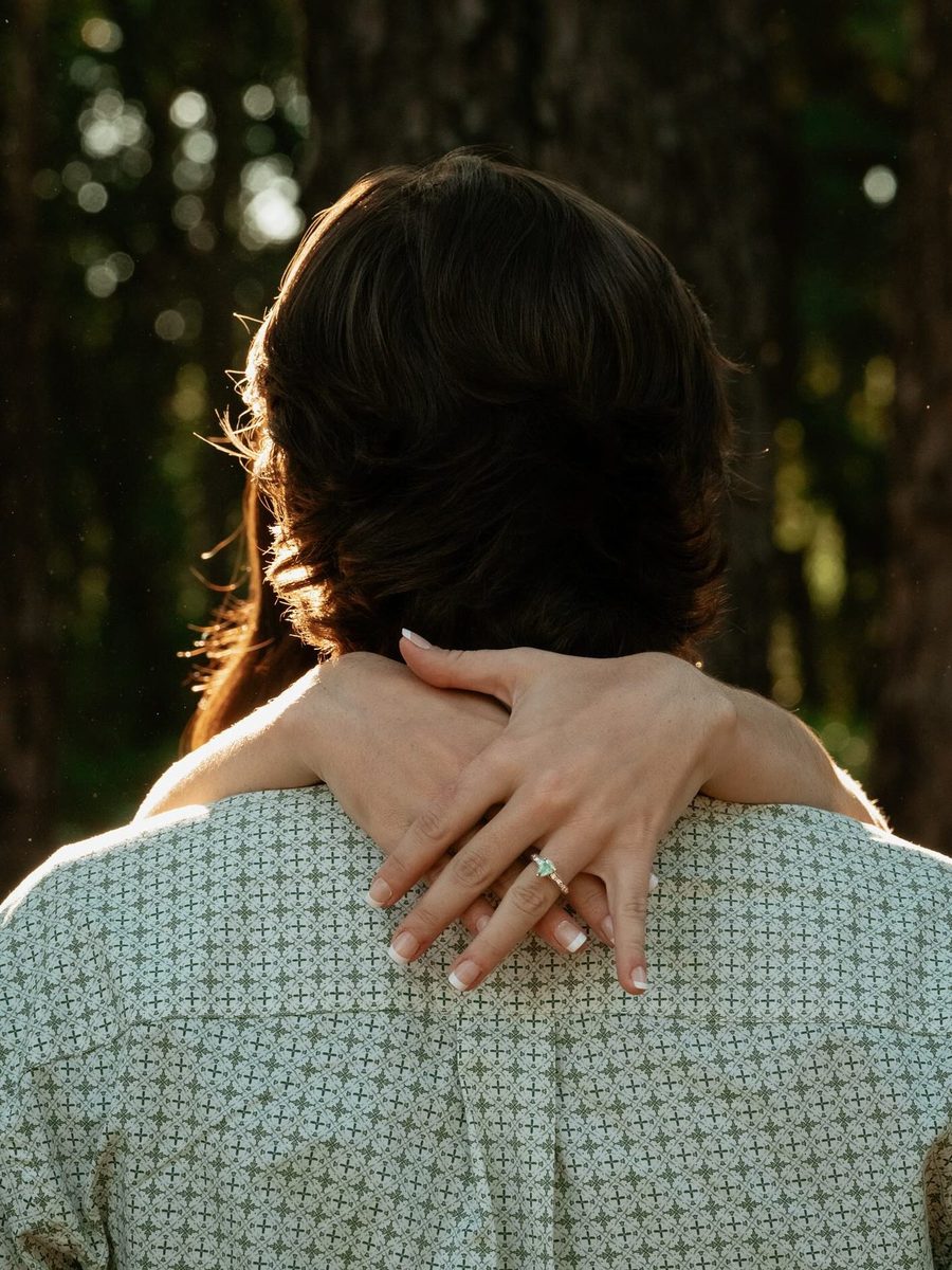 Engagement embrace from behind, ring resting on the shoulder, autumn woods beyond